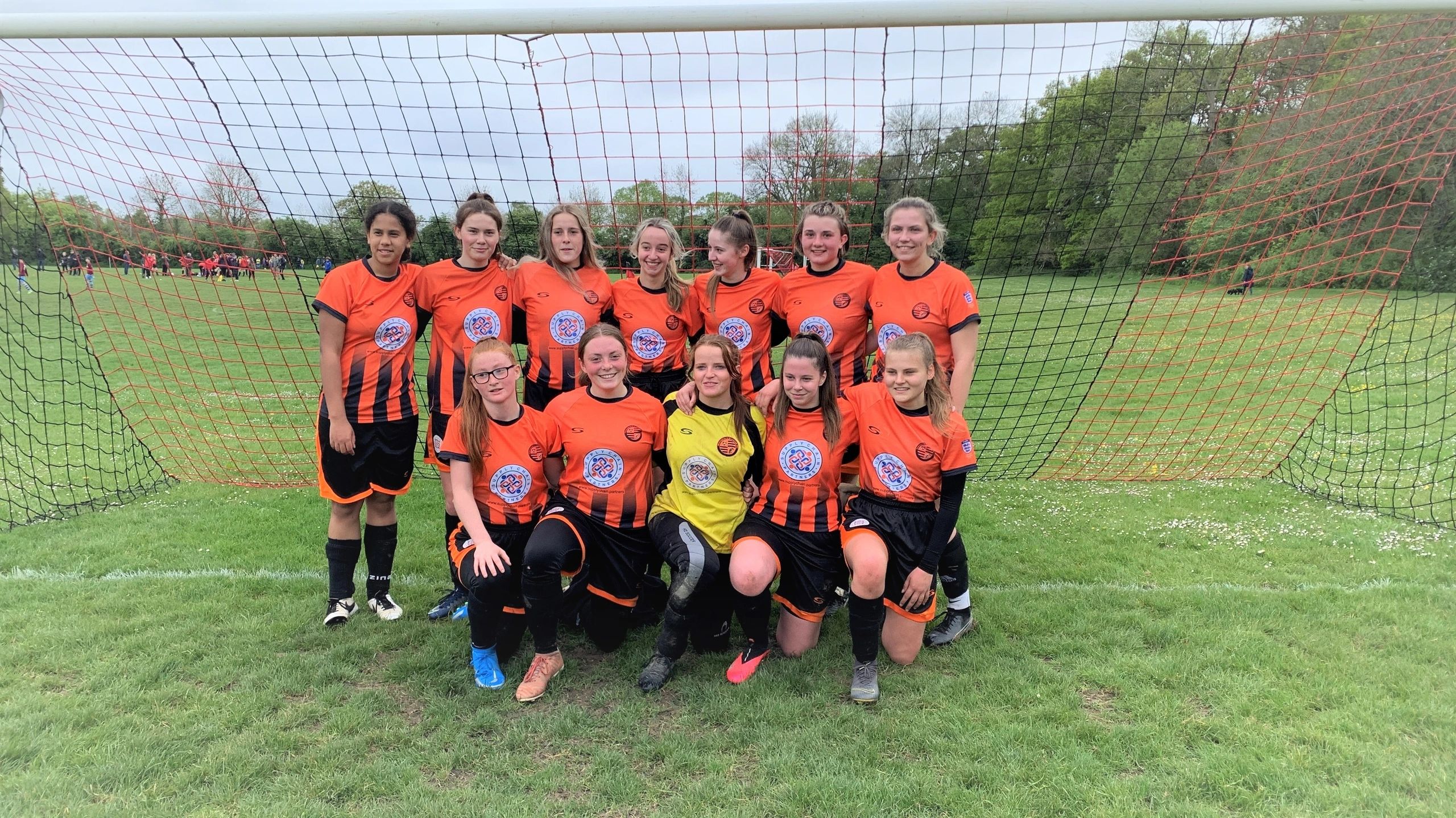 Wokingham and Emmbrook girls’ football team in orange and black shirts carrying the former SCP sponsor logo, posing in front of a goal.